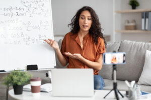 How to make math class interesting, Confident woman having video conference call using pc laptop. Female teacher showing whiteboard, recording tutorial with a mobile phone on a tripod, streaming live explaining math formula